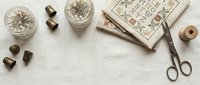 Vintage sewing supplies with antique scissors, wooden spools, glass buttons and embroidery pattern book on neutral fabric background for crafting projects.