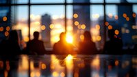 Blurred office interior with warm sunset light streaming through large windows creating golden bokeh effects and silhouettes of furniture against city skyline.