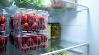 Fresh ripe strawberries in clear plastic containers stored in refrigerator with green vegetables and other healthy food items for meal preparation.