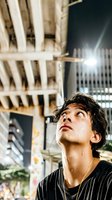 Young man with dark wavy hair and East Asian ethnicity gazing upward at illuminated urban skyscrapers at night, wearing black striped shirt, contemplative expression, city lights background.