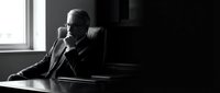 Senior male executive in formal business attire sitting at desk in modern office, speaking on telephone, contemplative expression, professional communication and corporate leadership concept.