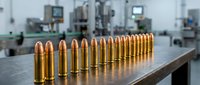Row of golden brass ammunition bullets lined up on metal table in modern manufacturing facility with industrial machinery in blurred background.