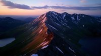 Majestic mountain peak illuminated by sunset glow, casting red-orange light on snowy ridges against purple sky, with alpine lakes nestled in valleys below.
