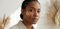 Young African American woman applying skincare product with fingers on face in natural light with pampas grass background for beauty and wellness concepts.