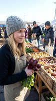 Young blonde woman in knit hat shopping for fresh vegetables at outdoor farmers market with wooden crates full of organic produce and other customers browsing.
