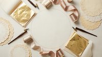 Elegant flat lay with gold leaf sheets, rose gold ribbon, lace doilies and calligraphy pens arranged around blank circular frame for luxury crafting projects.