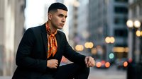 Young Hispanic businessman in black suit with orange patterned shirt sitting confidently in urban city environment with blurred lights background at dusk.