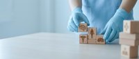 Healthcare professional in blue gloves arranging wooden blocks with medical icons on white surface for hospital management and patient care planning.