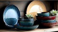 Handcrafted ceramic bowls and plates in turquoise blue and deep red glazes with rustic texture, stacked on wooden surface. Small succulent plants add natural accent.