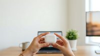 Hands holding white round jar with cream, lotion or skincare product on wooden desk with laptop and plant in background, suitable for beauty, cosmetics and wellness marketing.