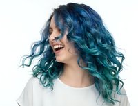 Young woman with vibrant blue-green curly hair laughing joyfully in white shirt on light background. Hair coloring and styling concept.