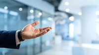 Businessman in dark suit extending open palm gesture in modern office environment with blurred glass partitions and bright lighting, conveying welcome, agreement, or presentation concept.