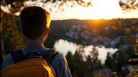 Young male hiker with backpack watching scenic sunset over lake and small town from hilltop viewpoint, back view silhouette in golden evening light.