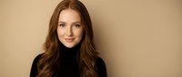 Professional young Caucasian woman with long wavy brown hair wearing black turtleneck sweater smiling confidently against neutral beige background.