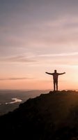 Silhouette of man with arms raised celebrating success on mountain peak at sunset with dramatic sky and valley landscape below.