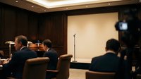 Male professionals in business suits seated in a conference room during a presentation or meeting, with projection screen and microphone visible for corporate training or seminar use.