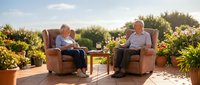 Senior couple relaxing on sunny terrace with wine, enjoying retirement lifestyle in comfortable armchairs surrounded by lush plants and garden setting.