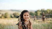 Young girl with braided hair and beaded accessories smiling outdoors in rural field setting, suitable for lifestyle, cultural diversity, and children's editorial content.