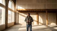Middle-aged Caucasian male construction worker standing in unfinished loft space with exposed brick walls and wooden beams during golden hour renovation project.