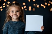Smiling young girl in gray sweater holding blank white paper against dark background with blurred golden bokeh lights, copy space for text or message.
