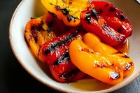 Grilled sweet bell peppers with charred skin in ceramic bowl, mix of red, yellow and orange peppers with smoky grill marks and natural oils on dark surface.