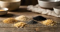 Assorted raw grains and seeds including quinoa, millet, chia and lentils arranged on rustic wooden table with ceramic bowls for healthy cooking recipes.