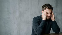Adult male with beard and dark hair, appearing stressed or fatigued, holding head in hands against neutral gray background. Suitable for mental health, workplace stress, anxiety, depression, and wellness content.