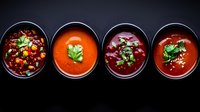 Fresh homemade soups and stews in black bowls on dark background, including tomato, beet and vegetable varieties garnished with herbs and sesame seeds.