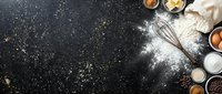 Baking ingredients arranged on dark surface with flour dusting and whisk, overhead view for culinary and recipe content.