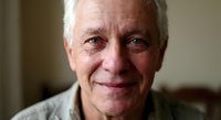 Senior Caucasian man with gray hair smiling warmly at camera in comfortable indoor setting, expressing contentment and wisdom through gentle facial expression.