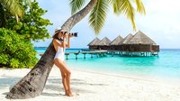 Woman in white swimwear leaning against palm tree on tropical beach, photographing overwater bungalows and turquoise lagoon. Travel photography concept.