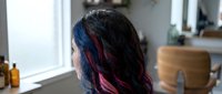 Young woman with long dark hair featuring vibrant blue and pink highlights, viewed from behind in a bright bathroom setting with natural window light and cosmetic products.
