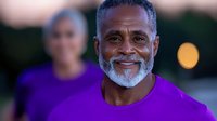 Mature African American man with gray beard wearing purple t-shirt smiling confidently at camera, with soft bokeh background and female figure in distance.
