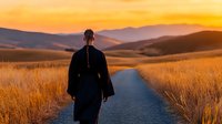 Young woman in long black coat walking alone on empty road through golden wheat fields at sunset, dramatic mountain landscape and orange sky in background.