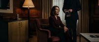 Professional businesswoman in burgundy suit sitting in luxury office interior with male colleague standing nearby for corporate meetings and workplace concepts.