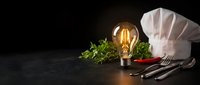 Culinary concept with glowing vintage light bulb, chef hat, fresh herbs and kitchen utensils on dark wooden surface for restaurant branding and cooking inspiration.