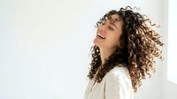 Joyful young Hispanic woman with curly hair laughing against white background, radiating happiness and positive energy in casual style.