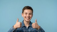Young Caucasian man in denim shirt showing double thumbs up gesture with bright smile against blue background for positive marketing campaigns.