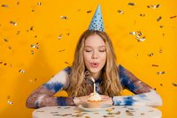 Young blonde woman blowing out candle on birthday cupcake with party hat against bright yellow background with falling confetti celebrating.