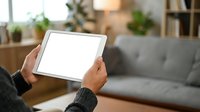 Person holding tablet with blank white screen in modern living room interior with sofa and plants for app mockup and digital display presentations.