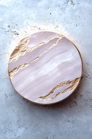 Luxurious round marble plate with gold kintsugi-style cracks on pale pink surface against textured gray background. Elegant tableware for modern dining.