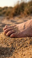 Caucasian child's foot partially buried in warm sand at beach during golden hour sunset, with dunes and beach grass blurred in background. Vacation, leisure, summer.