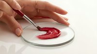 Close-up of female hands applying red cosmetic product on glass petri dish with brush, beauty formula testing concept.
