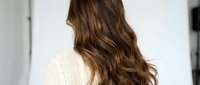 Young Caucasian woman with long wavy brown hair wearing cream knit sweater in modern interior, back view showing healthy shiny hair texture and styling.