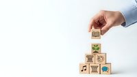 Hand placing wooden cube with brain icon on stack of blocks with various symbols representing skills and knowledge development on white background.