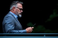 Mature businessman with gray beard and glasses in suit jacket checking smartphone outdoors against dark background with bokeh effect.