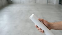 Male hand holding rolled white architectural blueprint or construction plan against blurred concrete interior background, suitable for construction, engineering, and business projects.