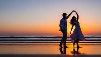 Romantic couple dancing on beach at sunset with golden sky and ocean waves reflecting on wet sand creating silhouettes for wedding and love concepts.