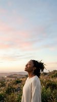 Woman with dark braided hair wearing cream cardigan gazes upward at golden sunset sky over rolling countryside landscape. Outdoor portrait for wellness, lifestyle, and inspirational content.