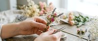 Florist hands trimming delicate dried flowers with scissors on rustic wooden table, creating a small bouquet with roses and lavender.